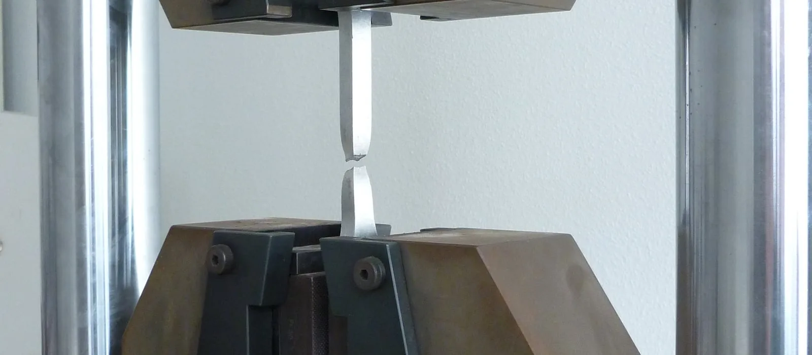 Corpo de prova chato rompido, ainda preso às garras de uma máquina universal de ensaios, com a estricção visível na região da fratura.