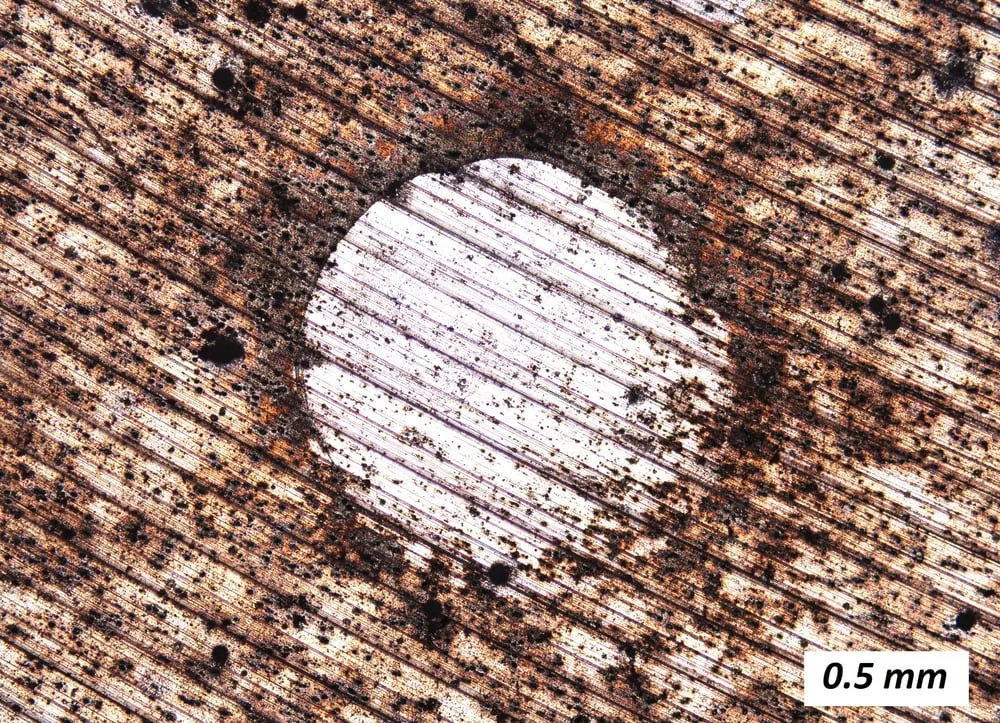 Micrografia óptica de superfície de duralumínio com pites escuros e produtos de corrosão, após imersão em água salgada.