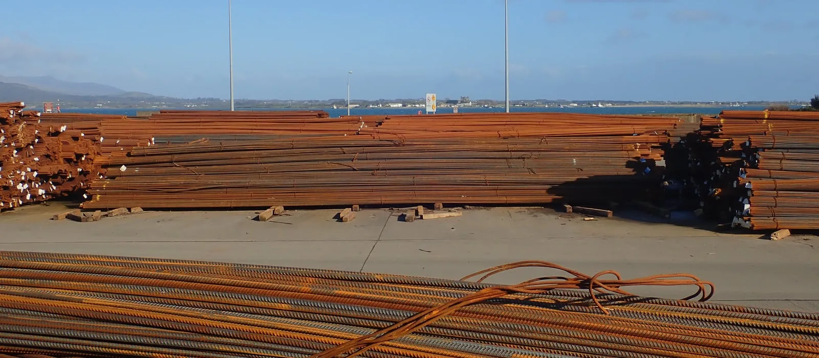 Feixes de barras de aço empilhados em um pátio portuário, com a superfície tomada por oxidação superficial.