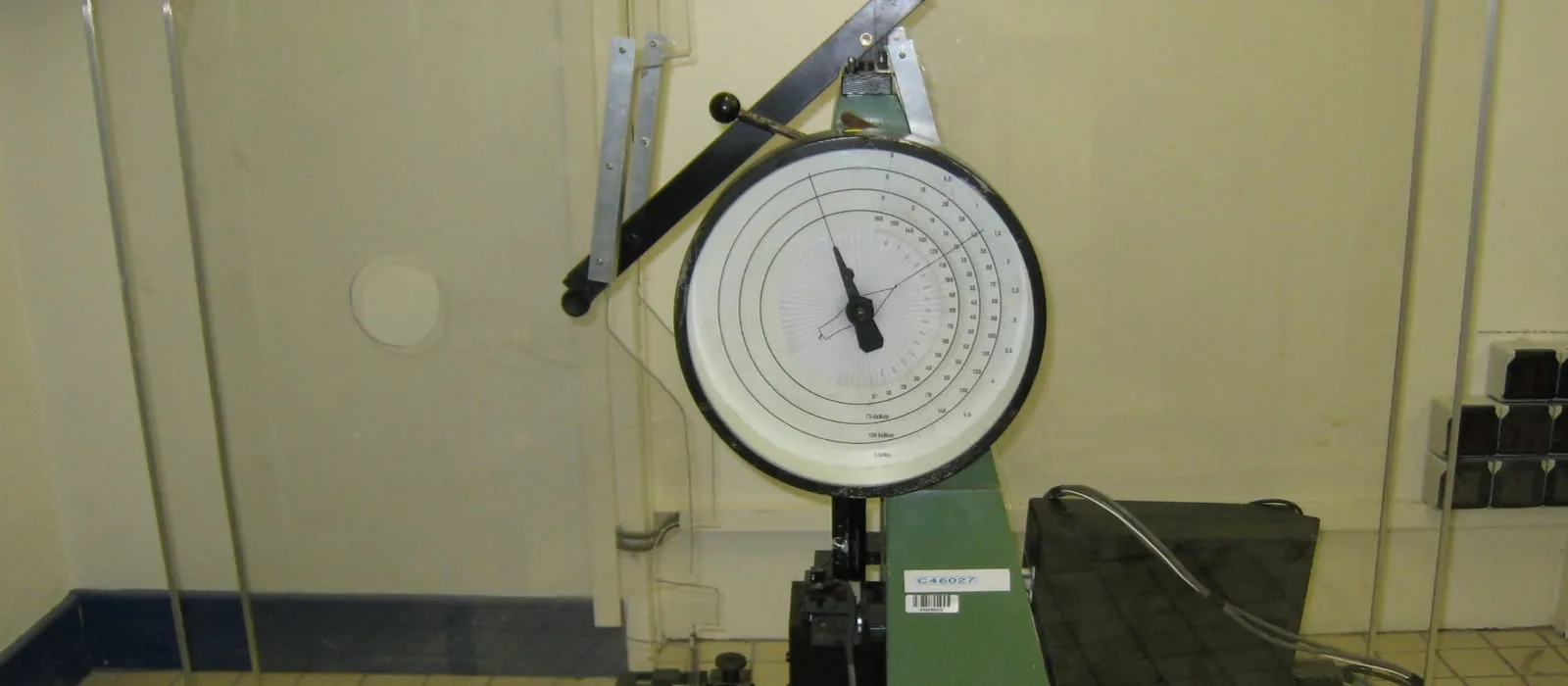Máquina de ensaio de impacto Charpy com pêndulo e mostrador analógico, em laboratório, com corpos de prova ao lado.